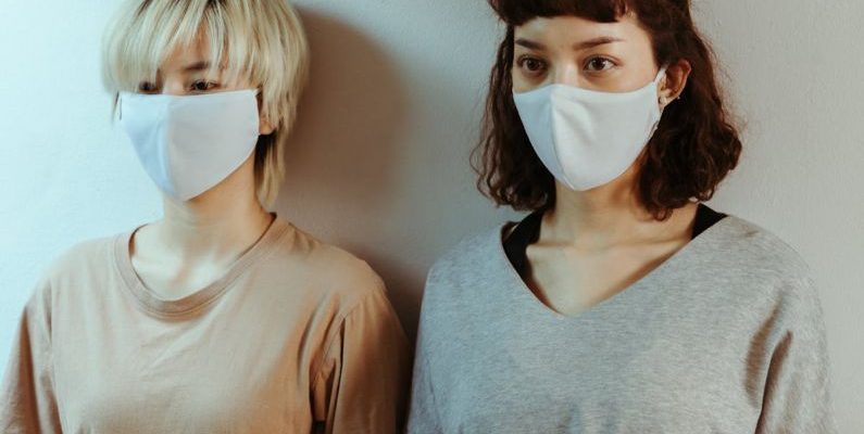 Benefits Of Staying - Asian women in home wear in medical masks attentively looking away while standing against white wall